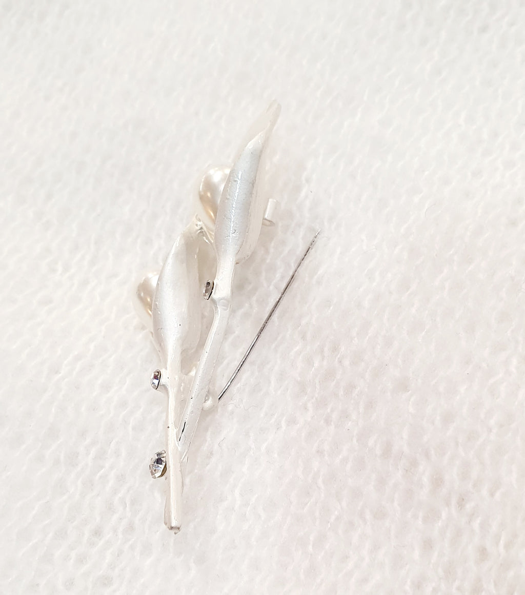 De Calliane is een verfijnde speldbroche geïnspireerd op de elegantie van de Calla-lelie.

De bloemvormige lijnen in zilver en wit worden verfijnd afgewerkt met kunststof parels en heldere kristallen, die het licht subtiel vangen.
Haar tijdloze ontwerp straalt rust en stijl uit — perfect voor sjaals, blazers of feestelijke gelegenheden.